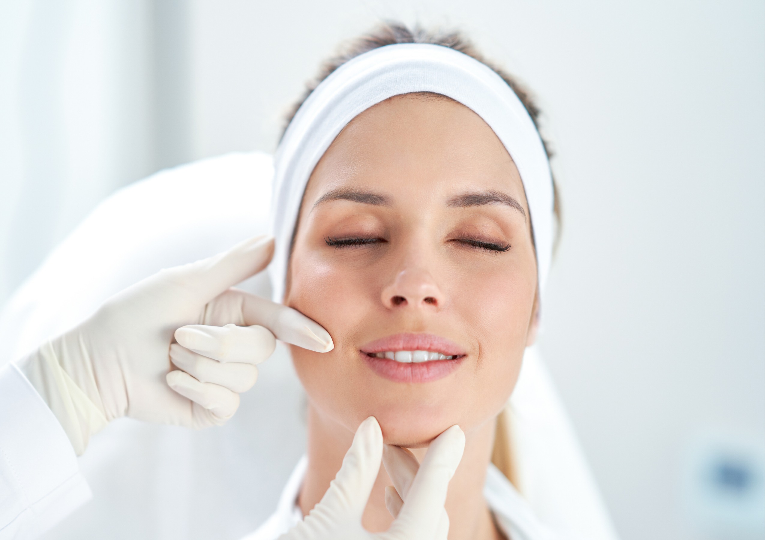 Close-up of a woman with a white headband and eyes closed, while a gloved practitioner gently lifts her chin for a facial treatment.